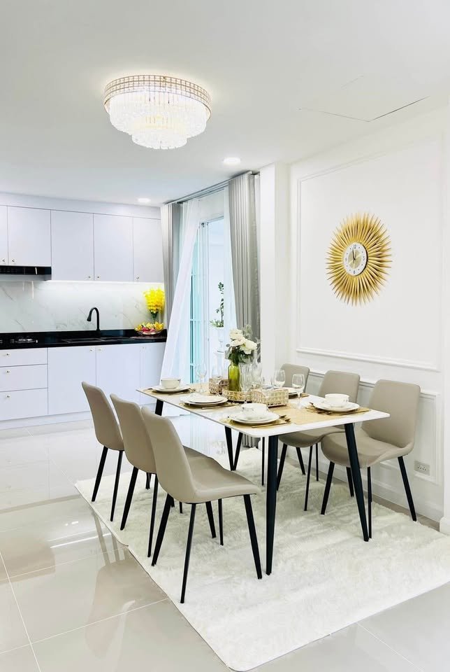 Bright open-plan dining area with a set marble table, six taupe chairs, crystal chandelier, gold sunburst wall clock, and white kitchen in the background