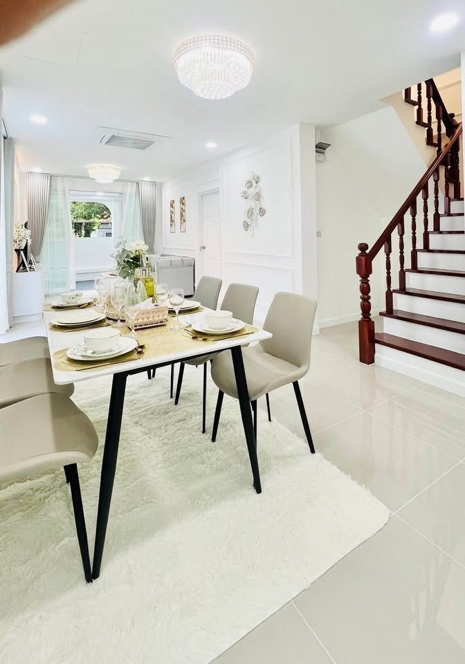 Bright dining area with a set table, taupe chairs, white fluffy rug, crystal chandelier, and a dark wood staircase nearby