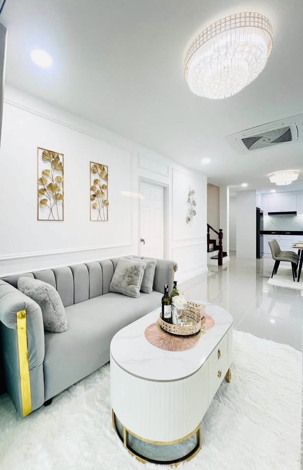 Elegant living room with a gray tufted sofa, white and gold oval coffee table, crystal chandelier, and gold leaf wall art