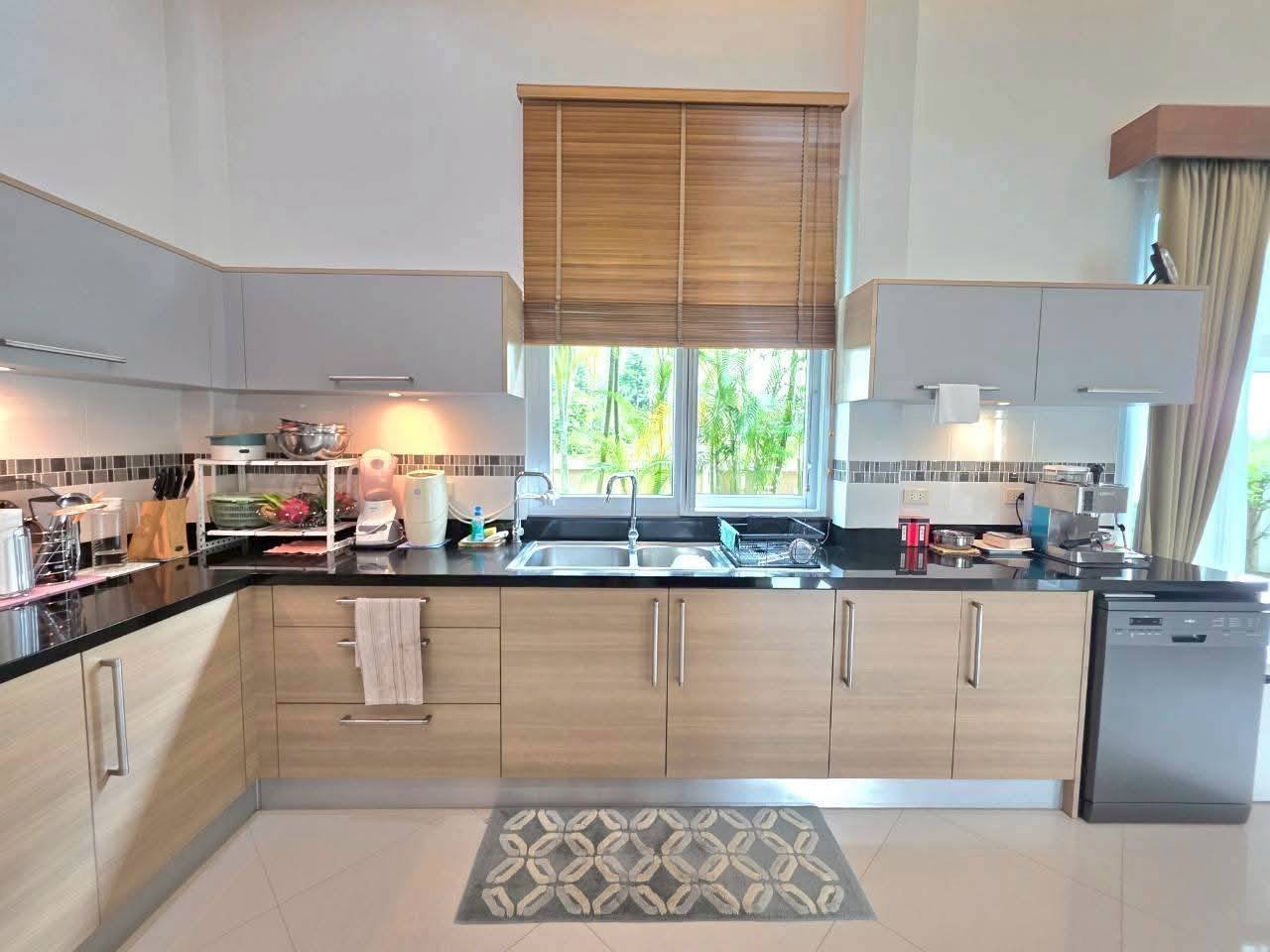 Spacious L-shaped kitchen with wood-finish cabinets, black countertops, double sink, dishwasher, and a window overlooking tropical greenery