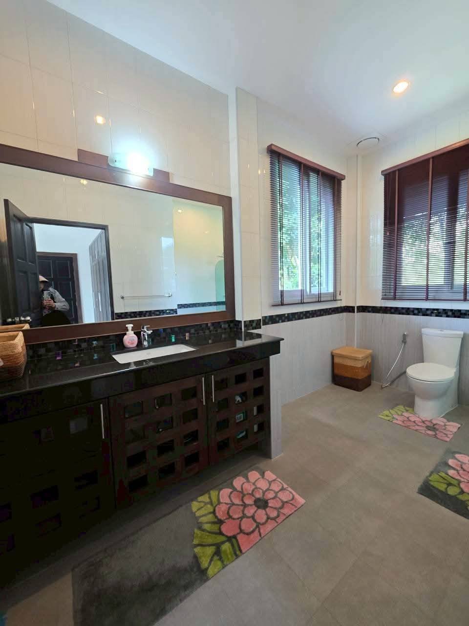Spacious bathroom with dark wood vanity, large mirror, toilet, floral bath mats, and two windows with blinds