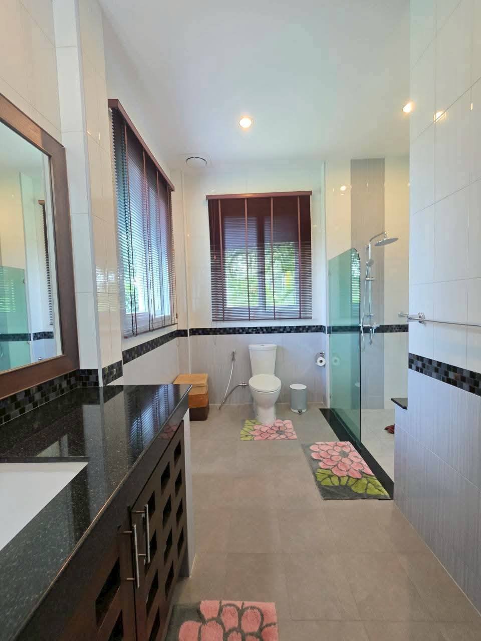 Spacious bathroom with dark granite vanity, glass-enclosed shower, toilet, and floral bath mats near windows with wood blinds