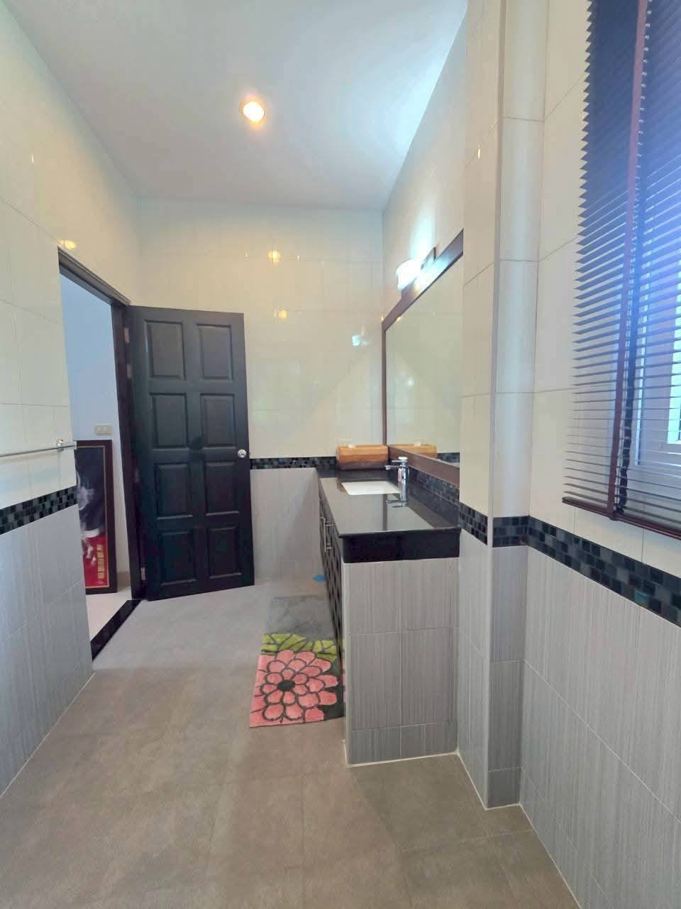 Spacious bathroom with dark countertop vanity, large mirror, tiled walls, wood blinds, and a floral bath mat on the floor