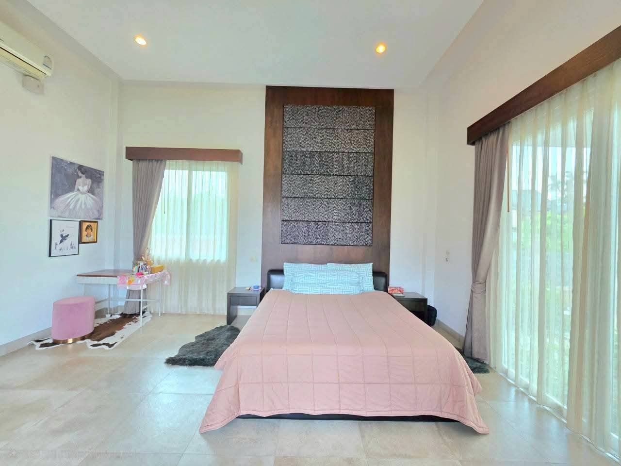 Spacious bedroom with a double bed, pink comforter, tall wooden headboard, sheer curtains, and a small vanity area with wall art