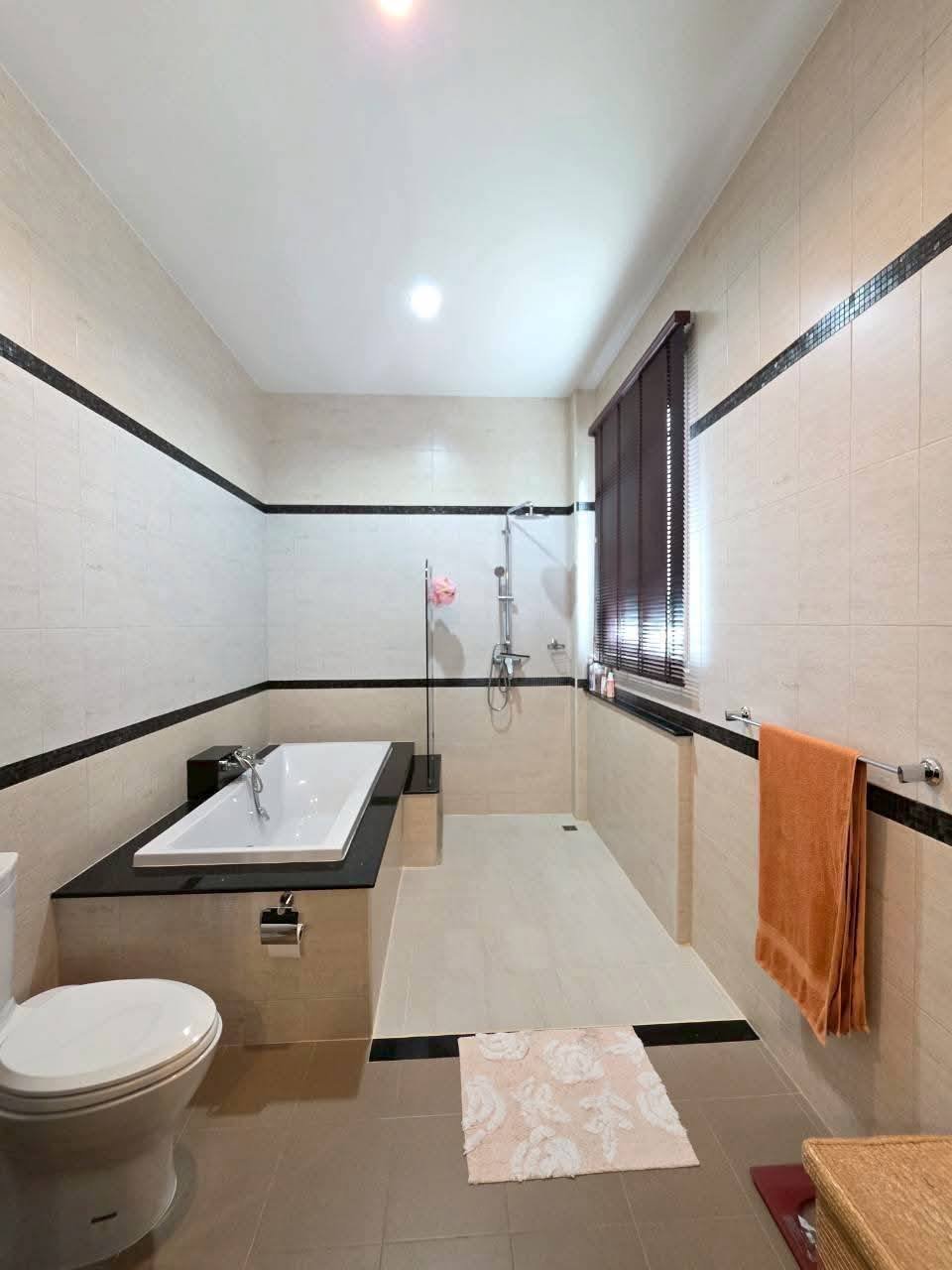 Spacious bathroom with beige tile walls, soaking tub on black surround, walk-in shower, toilet, and orange towel on rack