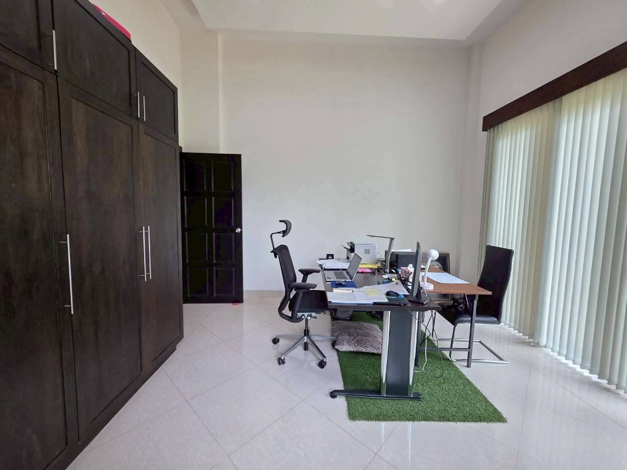 Home office with a desk, ergonomic chair, laptop, monitors, dark wood wardrobe, and vertical blinds on a large window