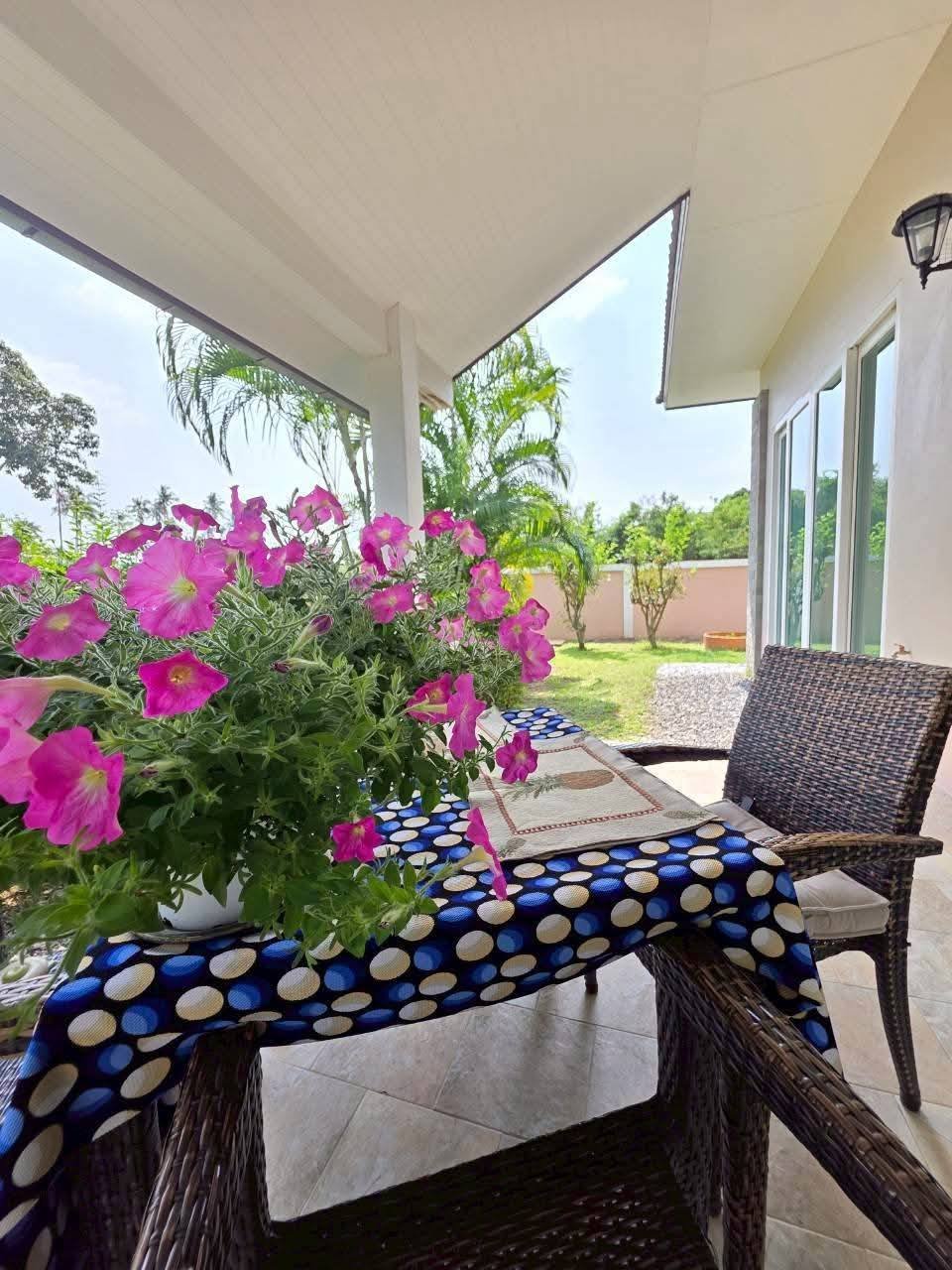 Covered patio with wicker chairs, a table with a polka-dot cloth, and a potted pink petunia plant overlooking a green garden