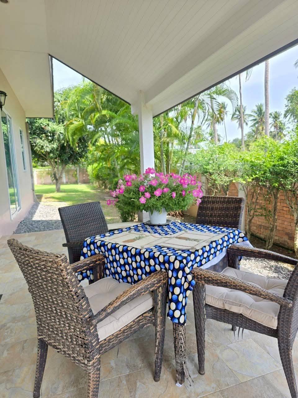 Covered patio with wicker chairs around a table with a blue polka-dot tablecloth and pink flowers, overlooking a lush garden