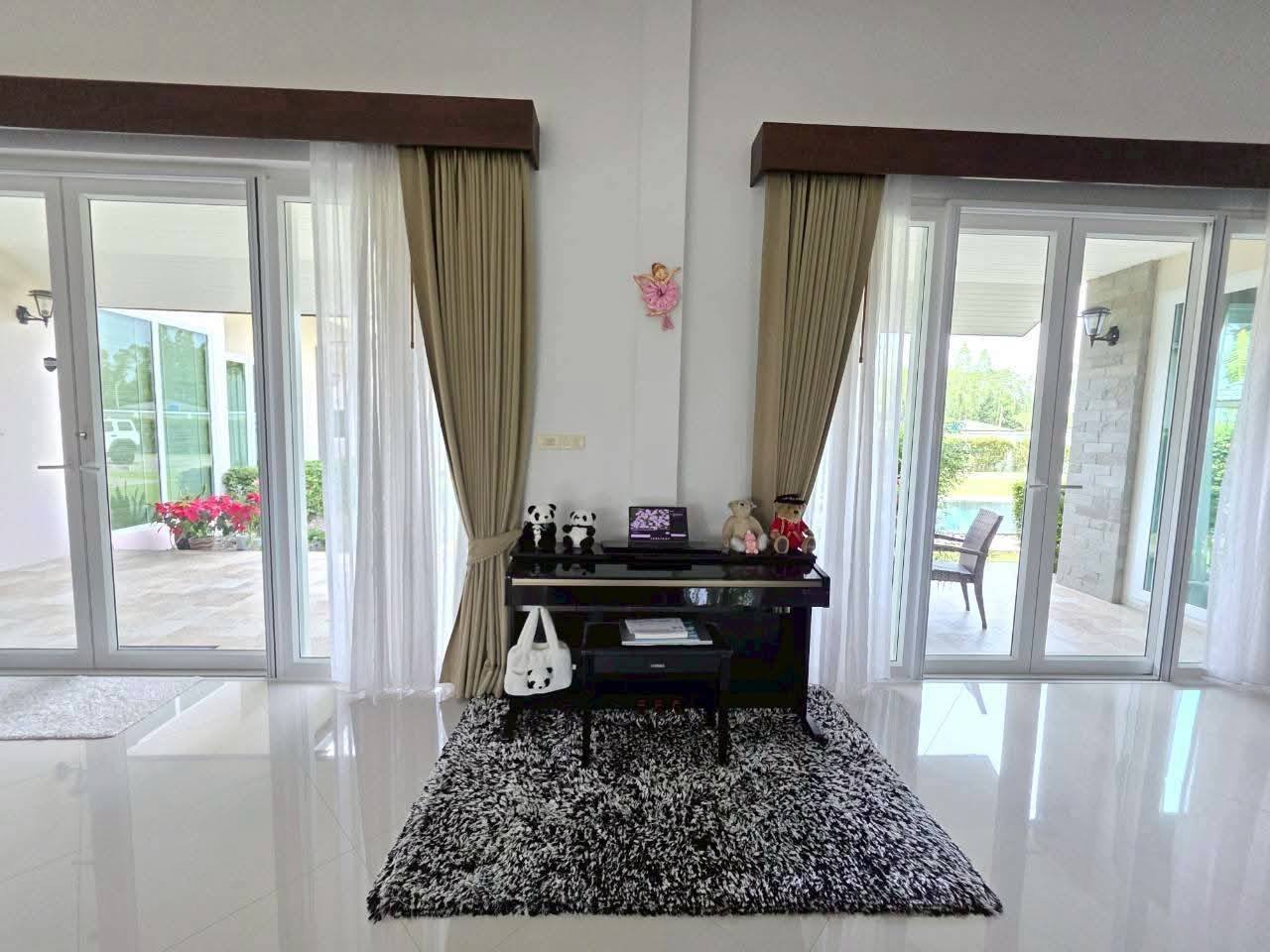 Bright living area with a black upright piano, stuffed animals on top, black-and-white shag rug, and large glass doors opening to a garden and pool patio