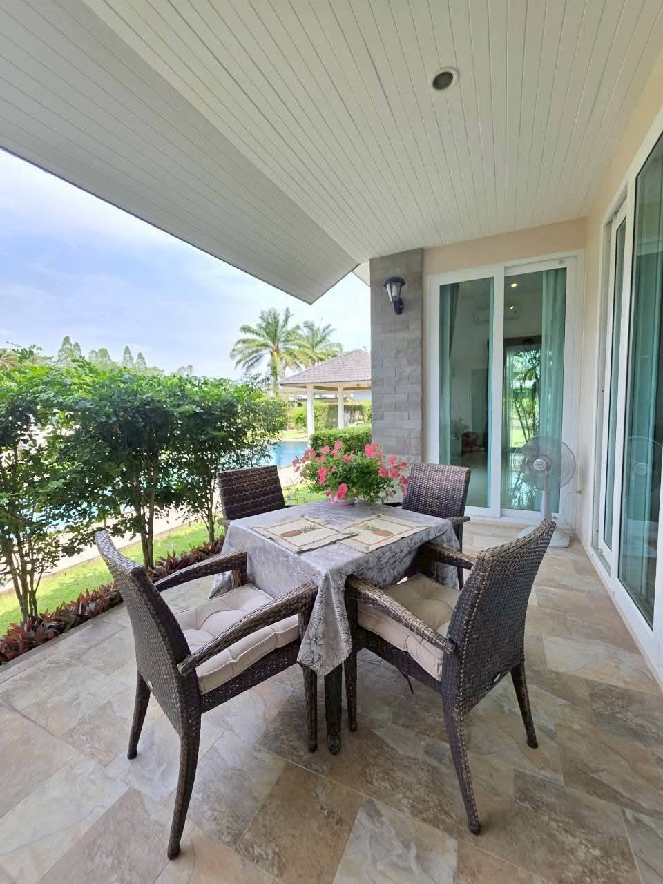 Covered patio with a wicker dining set for four, pink flowers on the table, overlooking a pool and tropical garden