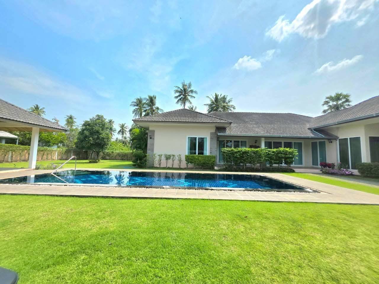 Single-story villa with a large outdoor swimming pool, manicured lawn, and tropical palm trees under a blue sky