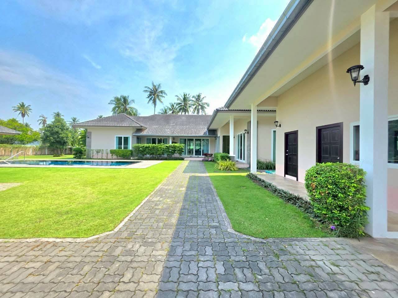 Exterior of a single-story villa with a paved pathway, manicured lawn, swimming pool, and palm trees in the background