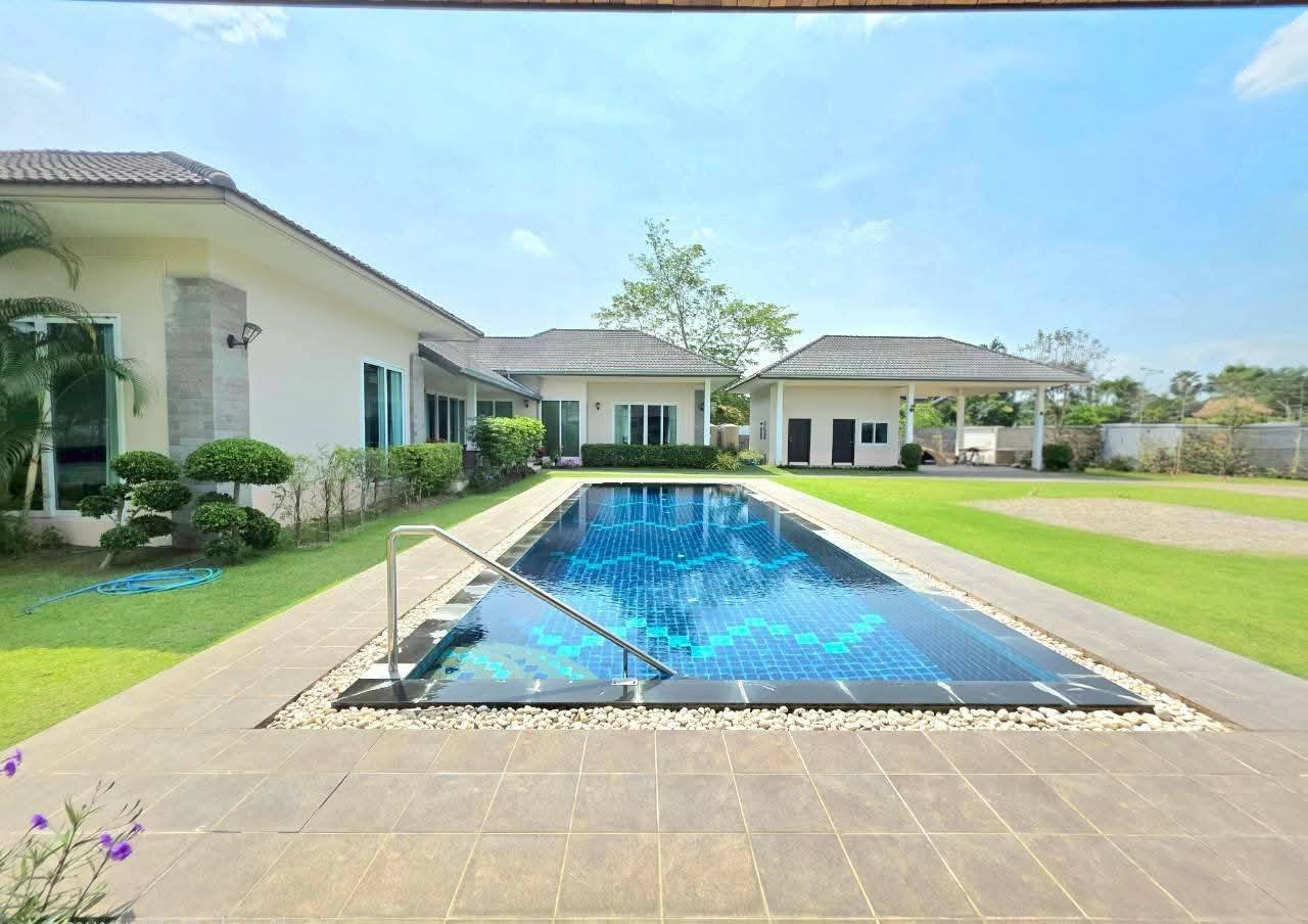Outdoor swimming pool with blue mosaic tiles surrounded by a paved deck, manicured lawn, and single-story white villa with guest house