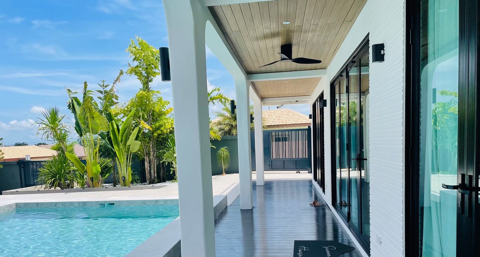 Covered outdoor patio with ceiling fan alongside a swimming pool, surrounded by tropical landscaping and glass-door facade