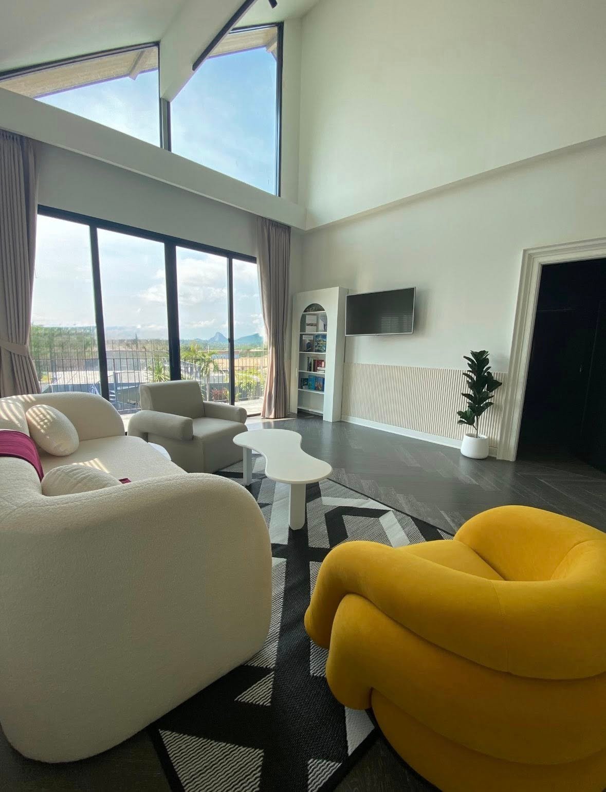 Bright modern living room with high ceilings, floor-to-ceiling windows with mountain views, white and yellow curved sofas, and a wall-mounted TV