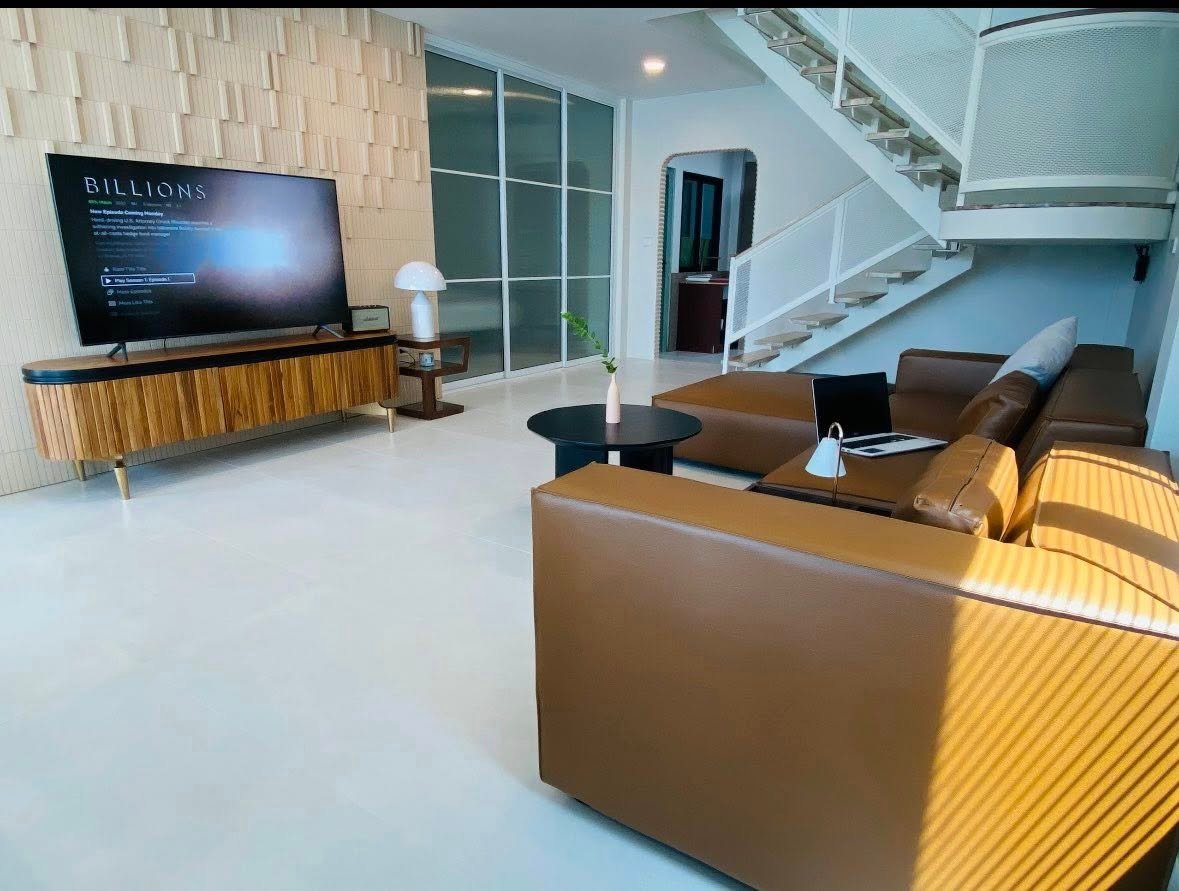 Modern living room with leather sofas, a large TV displaying Billions, a wood media console, and an open staircase