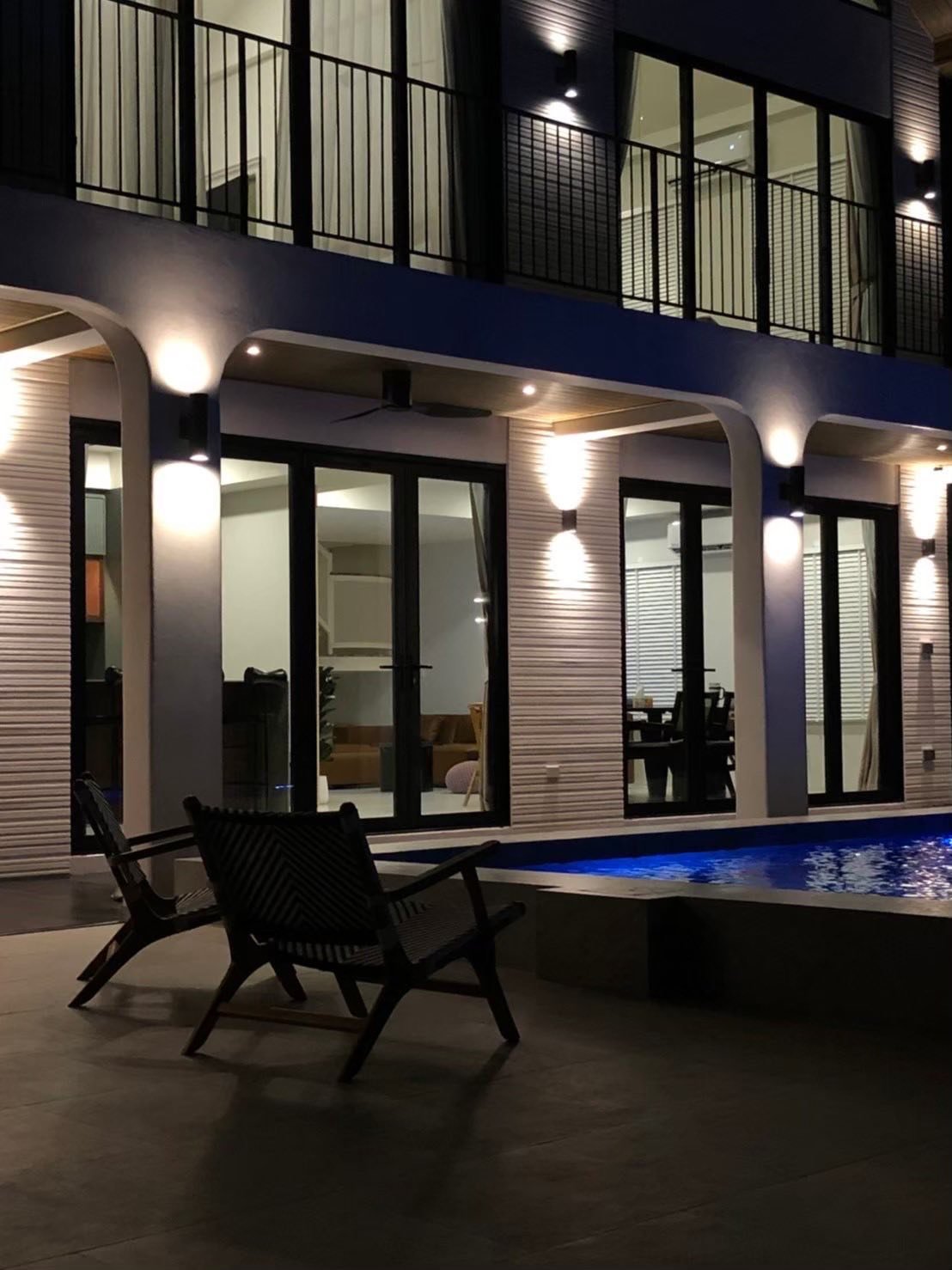Two lounge chairs beside a lit swimming pool at night, with a multi-story villa's illuminated exterior and glass doors in the background