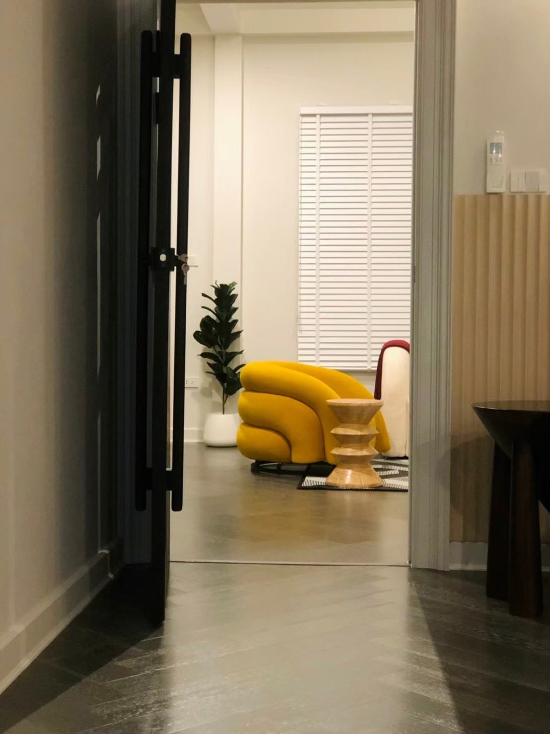 View through an open doorway into a bright room with a bold yellow curved armchair, wooden side table, and potted plant