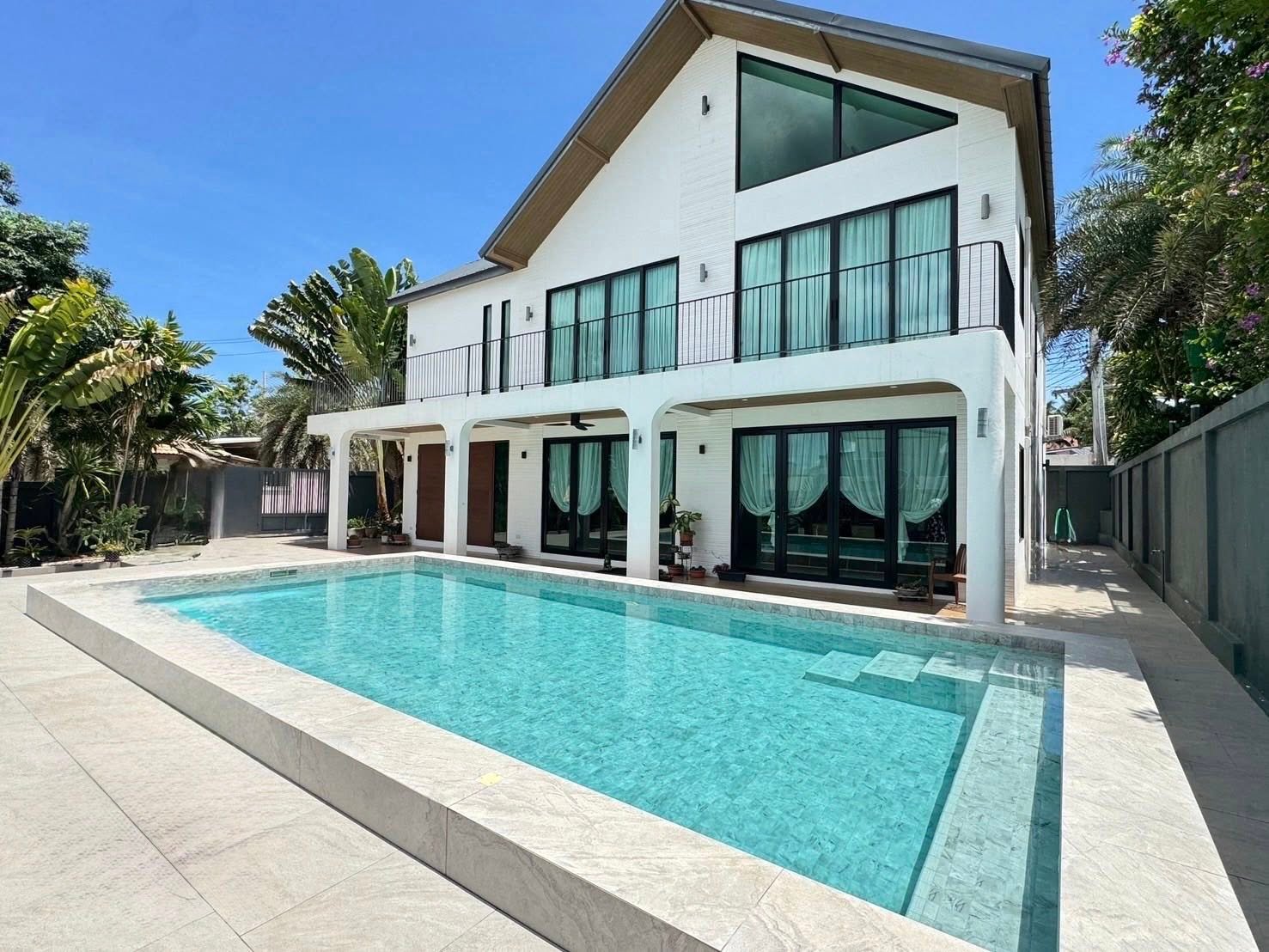 Two-story white modern house with large windows, balcony, arched patio, and a rectangular outdoor swimming pool surrounded by tropical trees