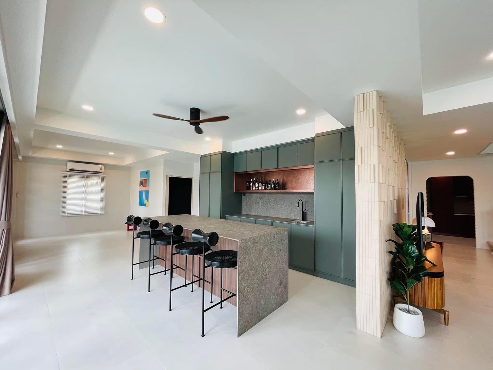 Open-plan living space with a stone-top kitchen island, black bar stools, sage green cabinetry, and a textured divider wall